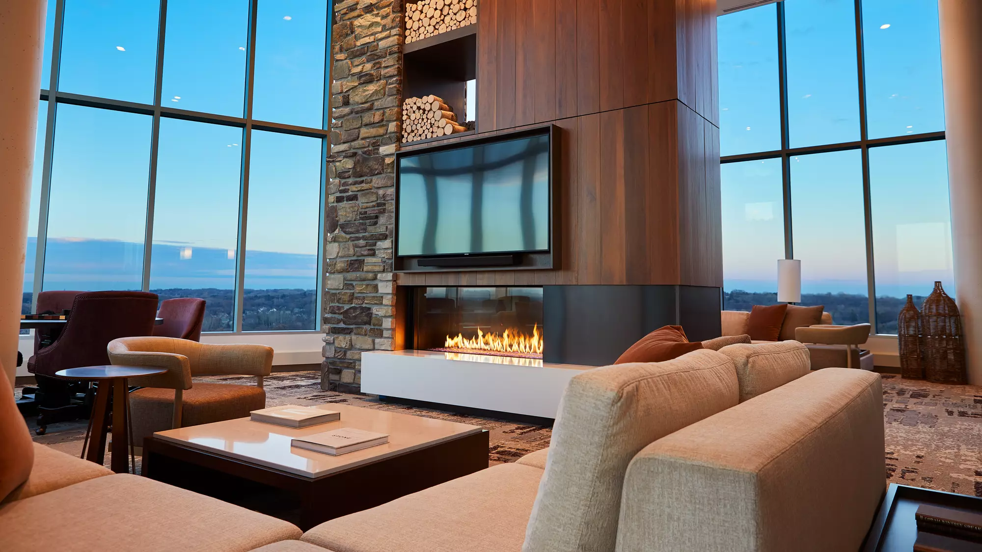A modern lounge with large windows offering a panoramic view, featuring an Enlight See-Through Gas Fireplace by Stellar by Heat & Glo with a mounted tv above, surrounded by comfortable seating and stylish decor.