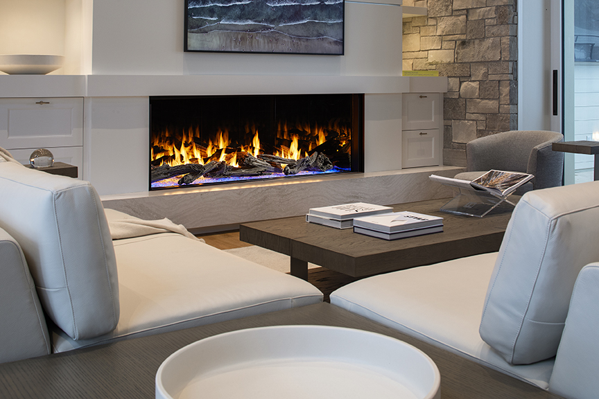 A stylish living room featuring two white sofas, a central wooden coffee table with books, and a modern, elongated electric fireplace set against a stone wall under a large painting.