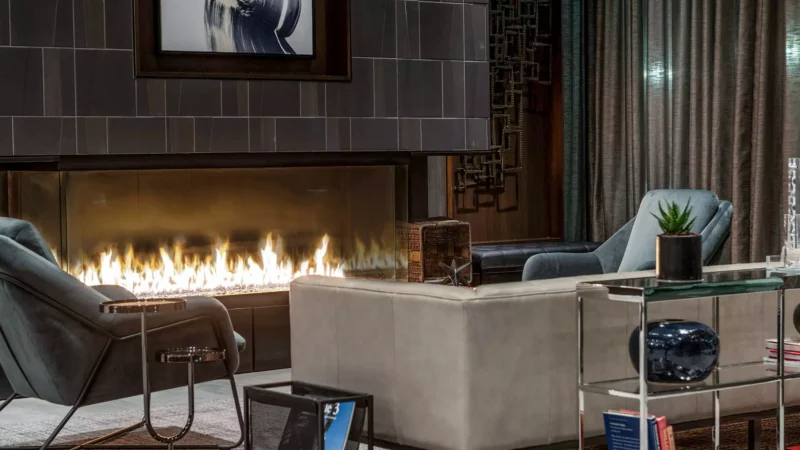 Elegant living room featuring the Enlight Bay Three-Sided Gas Fireplace by Stellar by Heat & Glo, surrounded by stylish furniture including a gray sofa, armchairs, and decorative items.