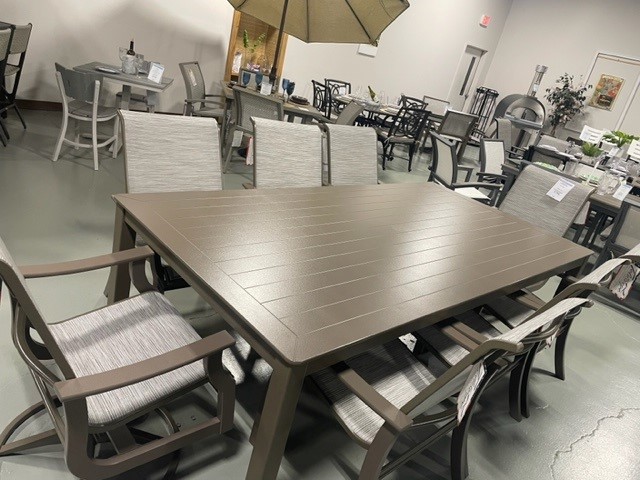 A showroom displaying various outdoor patio furniture sets, including St. Catherine Sling tables and chairs made from metal and fabric, arranged under an indoor setting with a concrete floor.