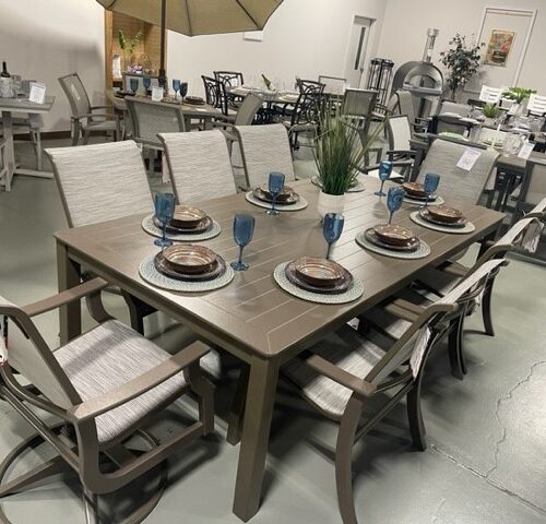 Furniture store display featuring a dining set with a rectangular table, six St. Catherine Sling chairs, and table settings including plates and blue glasses. Other dining sets are visible in the background.