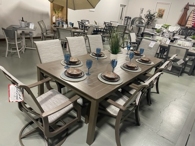 Furniture store display featuring a dining set with a rectangular table, six St. Catherine Sling chairs, and table settings including plates and blue glasses. Other dining sets are visible in the background.
