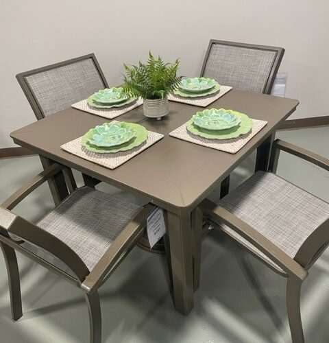 A modern Kendall Sling dining table set with four chairs, displayed with green placemats and plates, complemented by small potted plants as centerpieces in a light-colored room.