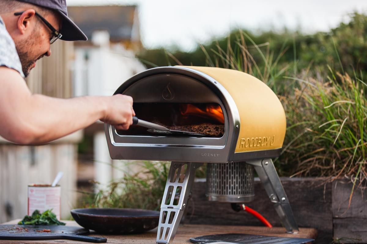 A man wearing a cap slides a pizza into a limited edition yellow Roccbox pizza oven placed on a wooden table outdoors, with plates and ingredients nearby.