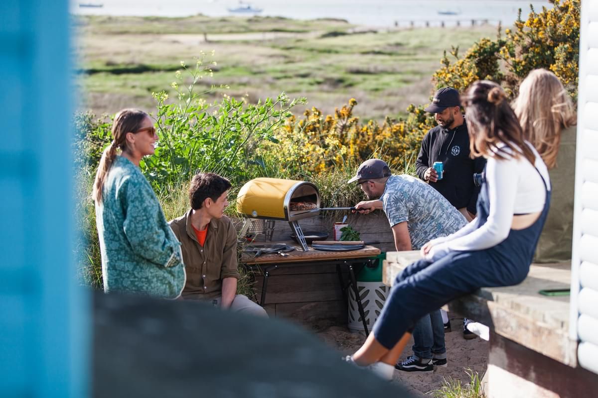 Group of people socializing outdoors near a limited edition yellow Roccbox pizza oven, with some engaged in conversation while one person attentively tends to the oven. They are surrounded by greenery and a
