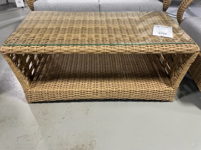 A rectangular wicker coffee table with a magnolia glass top and a shelf underneath, displayed in a showroom with a grey concrete floor. A price tag is visible on the table.