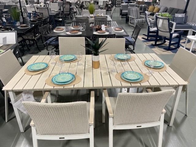 Modern Gensun Ventura outdoor dining set on display, featuring a light wood table and six beige wicker chairs, set with blue plates and glasses, in a showroom setting.
