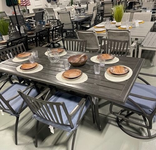 A dining area in a store with multiple tables and chairs displayed. The foreground shows a Gensun Amari wooden table set for four with plates, glasses, and silverware, surrounded by blue cushion