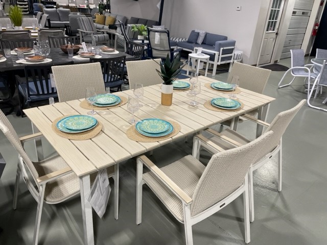 A modern Gensun Ventura outdoor dining set displayed in a showroom, featuring a light wooden table and six white woven chairs, each set with blue plates and clear glasses.