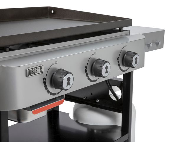 Close-up view of a Weber Griddle 28" showing three control knobs on its front panel, and a partial view of its flat top cooking surface and side shelf.