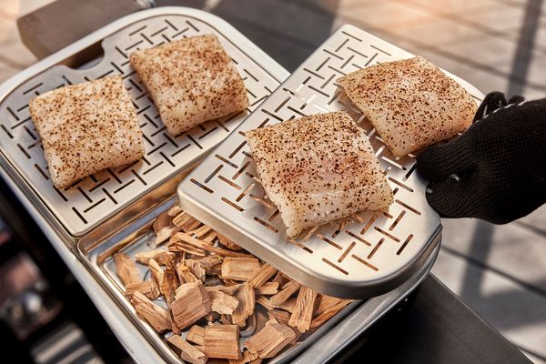 Four pieces of fish topped with spices are being smoked on a metal grill tray, with wood chips underneath. a hand with a black glove adjusts the tray.