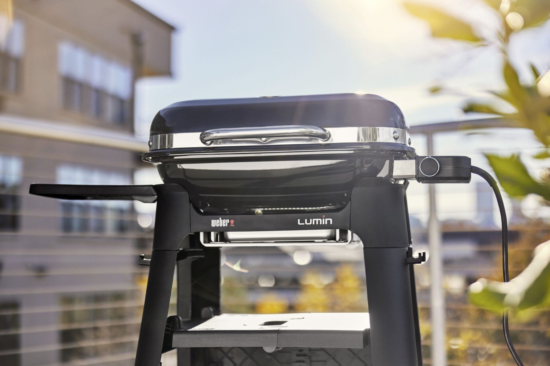 A modern weber lumin electric grill on a balcony, with a sunny backdrop casting a warm glow on its sleek, black and silver design.