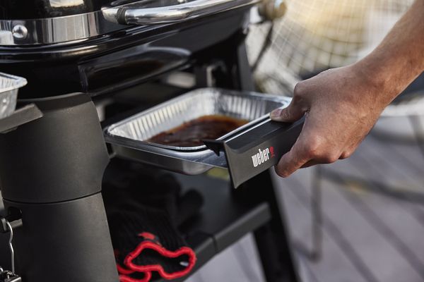 A person's hand pulls out a drip pan full of grease from a weber grill, with a focus on the clean details and the brand logo clearly visible.