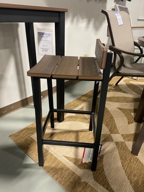 A modern Bazza bar stool with a dark wooden seat and black metal legs, displayed on a beige patterned rug in a showroom setting. Price tags are visible, indicating the stool is for sale.