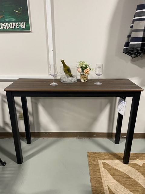 A wooden Bazza console table with black legs, displaying a wine bottle, two wine glasses, and a small floral arrangement, set against a neutral wall in a room with a gray floor.