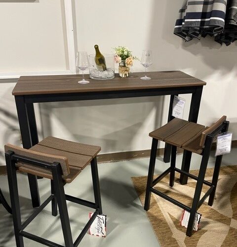 A modern dining setup with a narrow dark wood Bazza table and two matching stools, topped with a bottle of wine, two glasses, and a small floral arrangement in a brightly lit room.