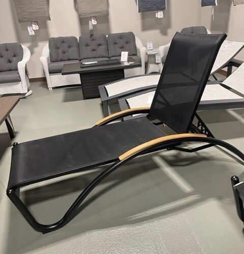 A modern black Helios Sling lounge chair with a mesh seat and wooden accents, displayed in a furniture store with various sofas and chairs in the background.