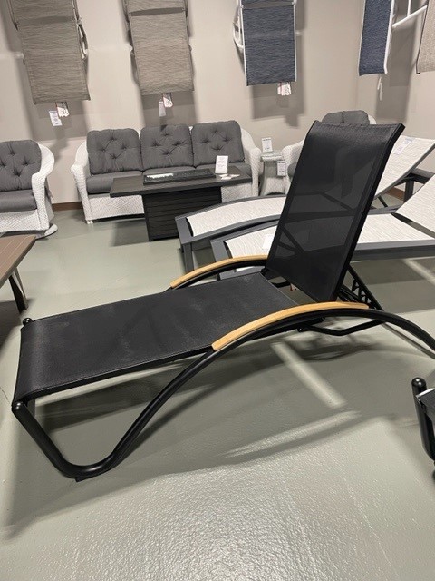 A modern black Helios Sling lounge chair with a mesh seat and wooden accents, displayed in a furniture store with various sofas and chairs in the background.