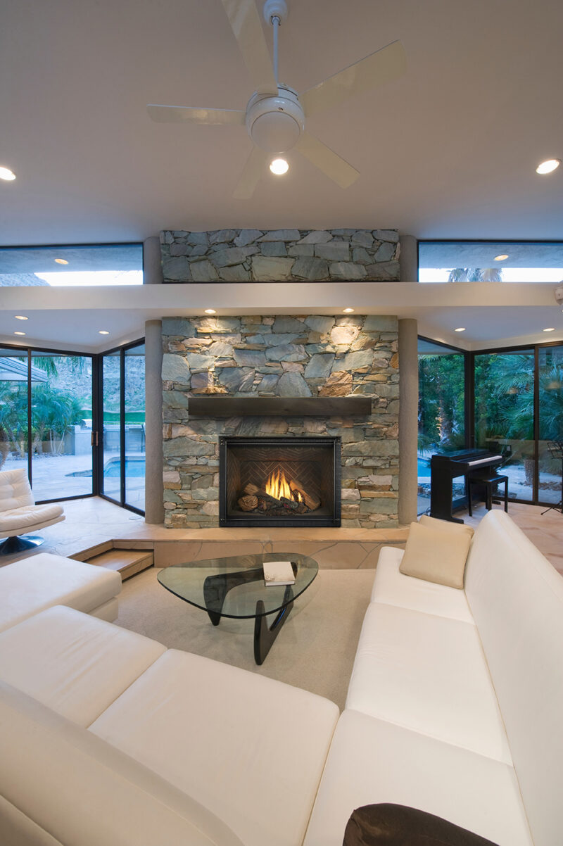 Modern living room with large white sectional sofa, stone fireplace, glass coffee table, and a ceiling fan. large windows provide a view of the outside.