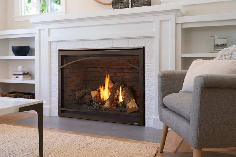 A cozy living room with a Heatilator Caliber Gas Fireplace, featuring realistic logs and flames. A grey sofa is partially visible on the right, with built-in white shelves in the background.