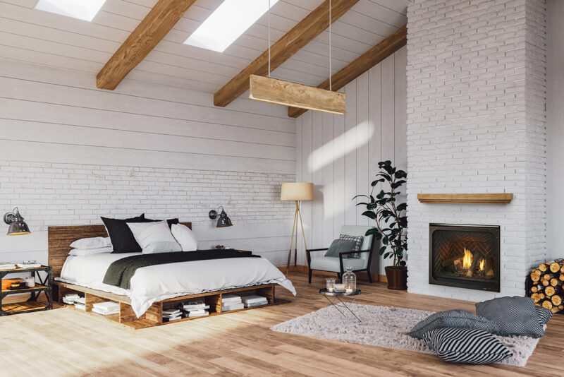 A cozy bedroom featuring a white brick wall, a wooden beamed ceiling, and a Heatilator Caliber Gas Fireplace. There is a plush bed, a floor lamp, an armchair, and