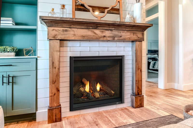 A cozy living room featuring a Heatilator Caliber Gas Fireplace with a rustic wooden mantel, surrounded by white shiplap and decorative light green cabinetry. A mirror and decorative items rest above