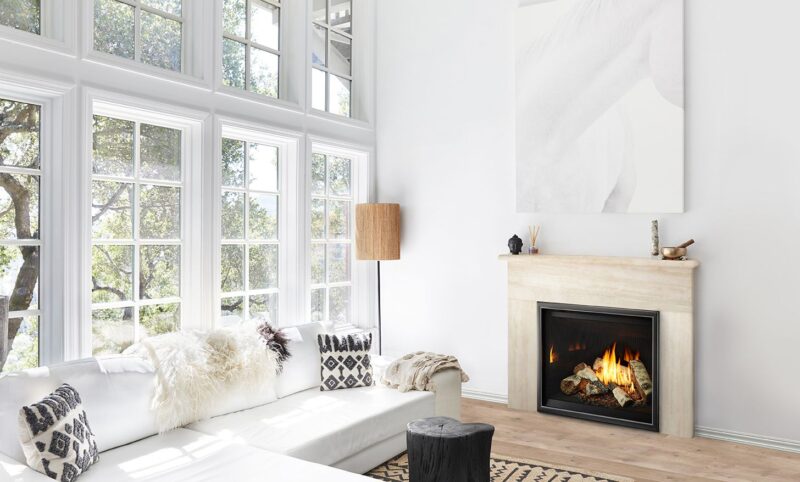 A bright and airy living room with large windows, a white sofa, patterned cushions, and a Heat & Glo 6K/8K Series Gas Fireplace. A small stool and a lamp beside