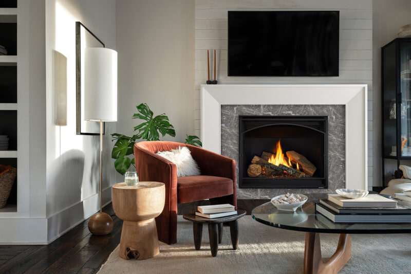 A cozy living room featuring a Heat & Glo 6K/8K Series Gas Fireplace, a red armchair with a fur throw, a white sofa, built-in bookshelves, and wooden