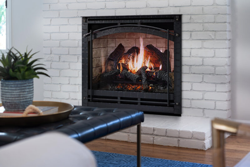 A cozy living room with a roaring SimpliFire Inception Electric Fireplace set into a white brick wall, framed by a black metal grate. The room is decorated with a plant, a blue rug,