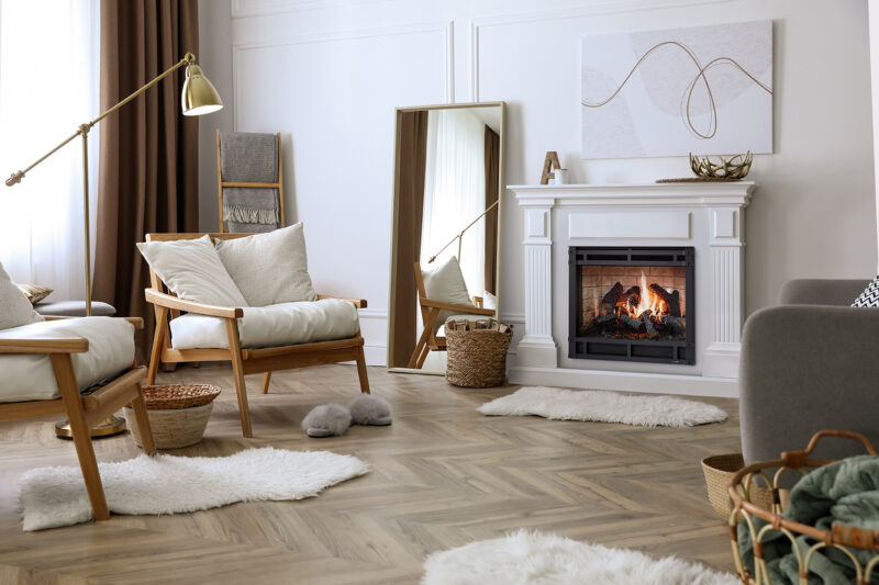 A cozy living room featuring a SimpliFire Inception Electric Fireplace, two beige armchairs, a large mirror leaning against the wall, art pieces, and various woven baskets and plush rugs.