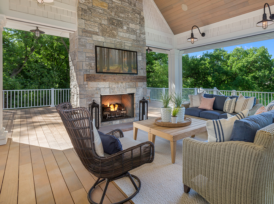 A cozy outdoor patio with a Vesper Outdoor Fireplace, wicker furniture, and wooden floor set against a background of lush green trees under a sunny sky.