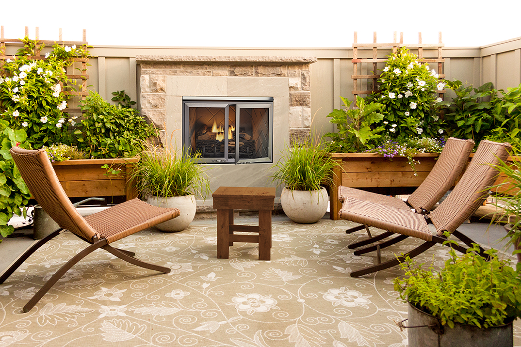 An inviting rooftop patio area with a central Vesper Outdoor Fireplace, surrounded by lush plants in wooden containers, two lounging chairs, and a small wooden table on a patterned rug.