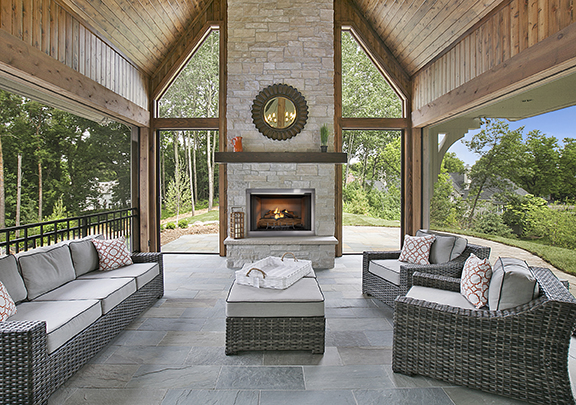 An elegant outdoor living space featuring wicker sofas with cushions, a Vesper outdoor fireplace, and a high wooden ceiling, surrounded by nature.