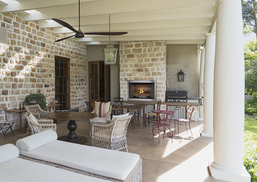 An elegant covered patio features wicker furniture, a round wooden table, a Vesper outdoor fireplace, and a built-in grill, surrounded by stone walls and white columns.