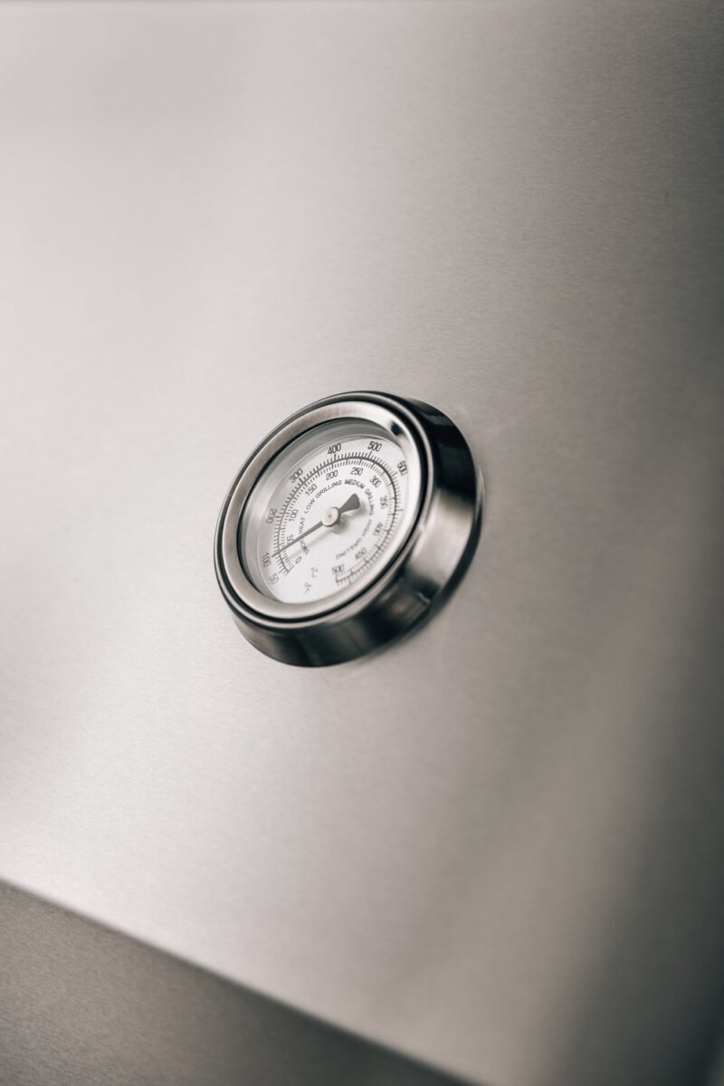 A close-up photo of a circular analog thermometer with a white dial, mounted on a metallic surface, showing a temperature reading. the background is softly lit and monochrome.