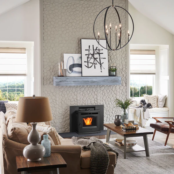 A cozy living room featuring a beige sofa, a wood stove with visible fire, abstract wall art, and modern decor under a large circular chandelier, surrounded by sunlit windows.