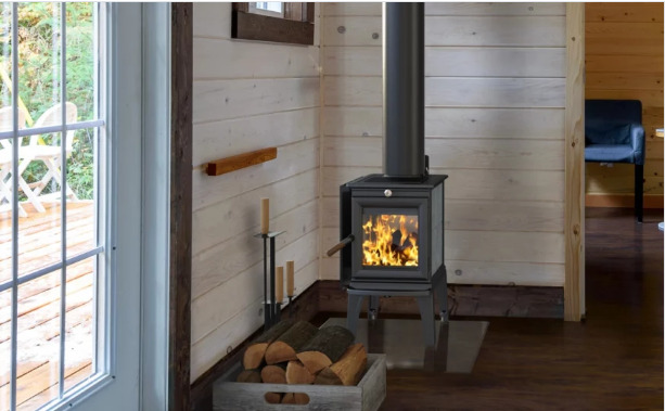 A cozy cabin interior featuring a lit wood stove with visible flames, a neatly stacked wood basket beside it, and light wooden walls that complement a bright outdoor view visible through a window.