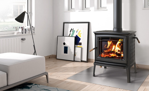 A cozy living room featuring a lit wood-burning stove, a minimalist white couch, and a floor lamp. art supplies and canvases lean against a wall near a large window.