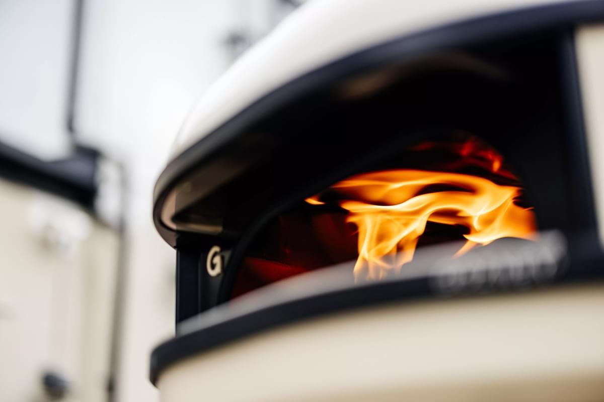 Close-up of vibrant flames inside a pizza oven, with the brand name partially visible on the front, set against a blurred building background.
