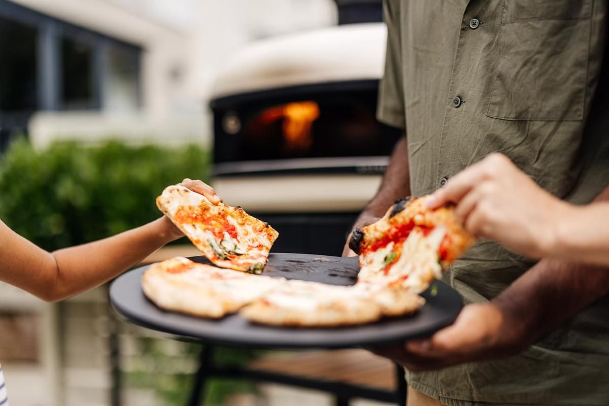A person in a green shirt serving slices of freshly cooked pizza from a black plate to another person, with a portable pizza oven in the background.