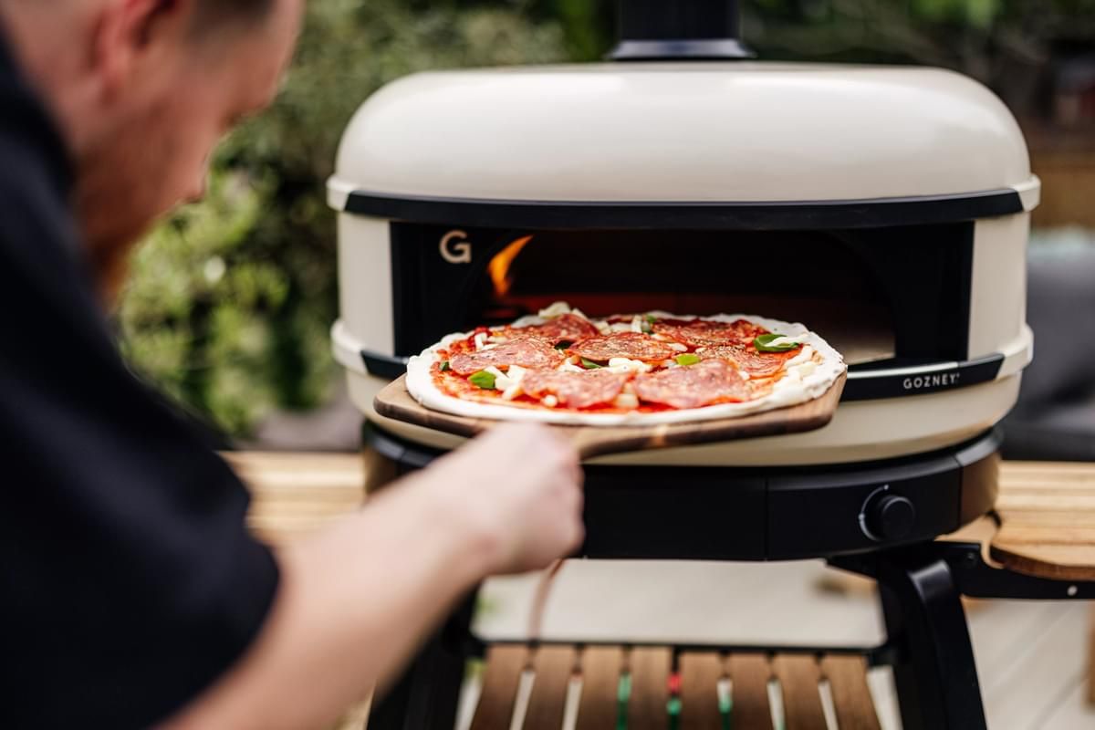 A person is sliding a pepperoni pizza into a portable outdoor pizza oven using a wooden peel. the focus is on the pizza and the oven, with the person visible from the back.