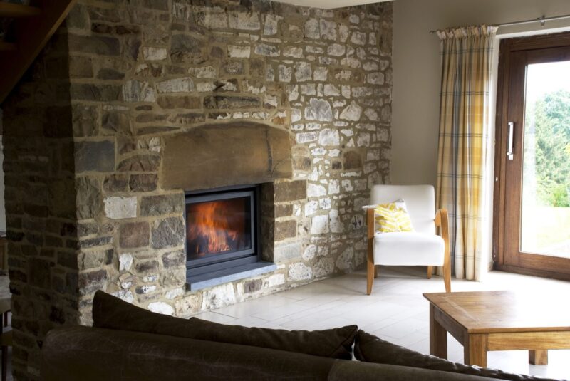 Cozy living room featuring a stone fireplace with a lit fire, a white armchair with a yellow cushion, and a wooden coffee table, adjacent to a glass door leading outside.