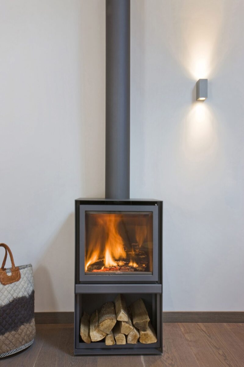A modern wood-burning stove with visible fire, placed in a room corner beside a beige wall, under a wall-mounted light. a storage space below the stove holds neatly stacked logs.