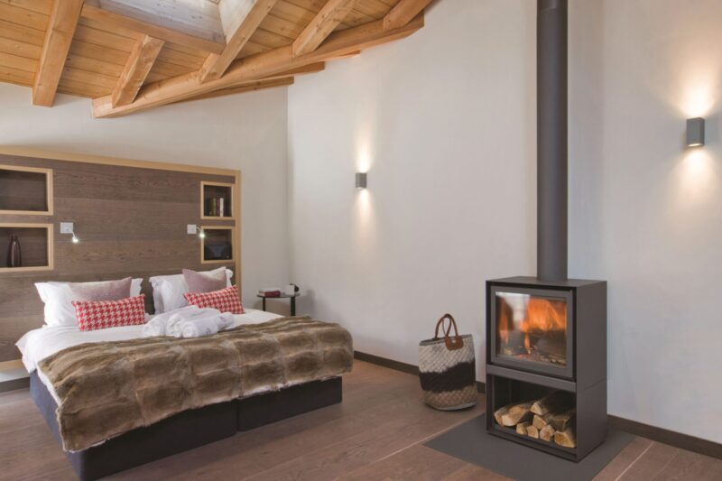 A cozy modern bedroom featuring a large bed with a brown blanket and red and white pillows, wooden ceiling beams, white walls, and a small freestanding fireplace.