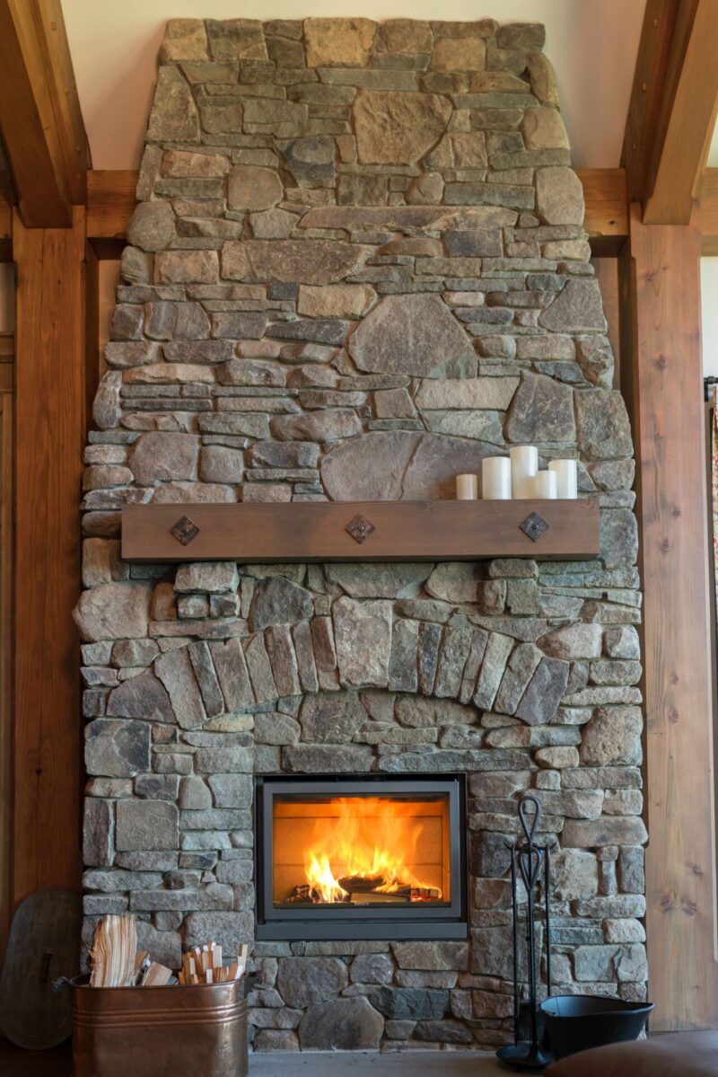 A cozy fireplace with a blazing fire, surrounded by a tall, rough stone chimney and wooden frame, with decorative items on the mantel.