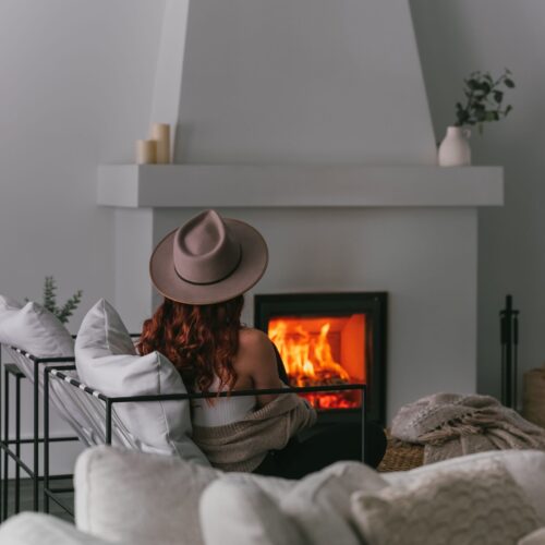 A woman with red hair, wearing a beige hat and sweater, sits facing a glowing fireplace in a cozy room with neutral-toned decor.