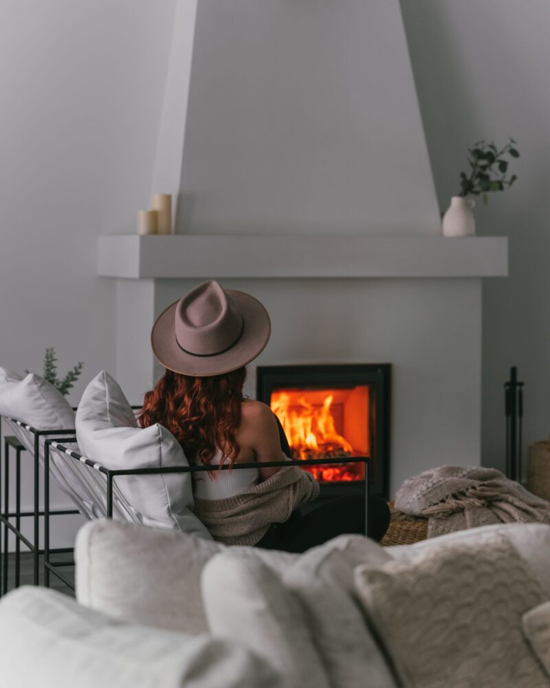 A woman with red hair, wearing a beige hat and sweater, sits facing a glowing fireplace in a cozy room with neutral-toned decor.