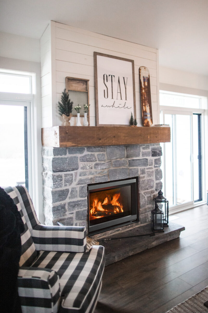 A cozy living room with a stone fireplace, on which a wooden mantle displays a "stay awhile" sign, flanked by small plants. a striped armchair and lantern are visible in the foreground.