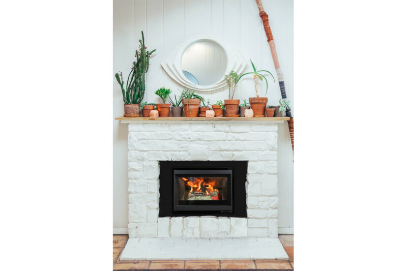 A cozy fireplace with a roaring fire, surrounded by a white brick mantel adorned with a circular mirror and several potted plants including cacti and succulents.