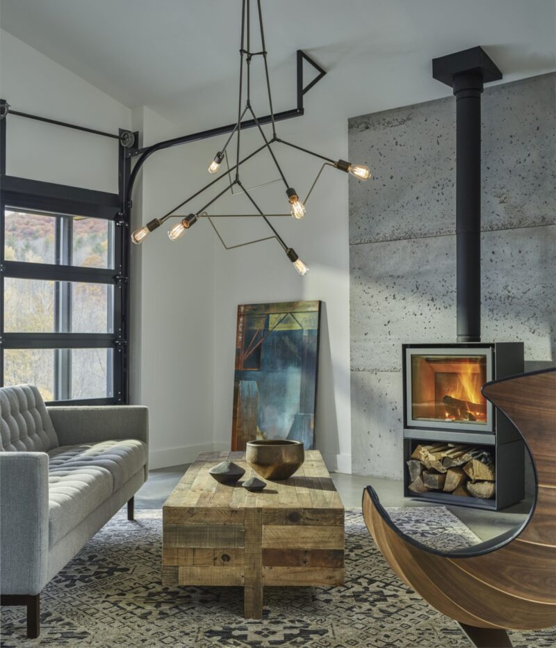 A modern living room featuring a large fireplace, gray sofa, geometric chandelier, and a rustic wooden coffee table. the room is accented by large windows and a unique curved chair.
